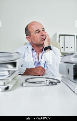Portrait auf nachdenkliche reife Arzt für Allgemeinmedizin in seinem Büro Tisch mit Binder und Ordner von medizinischen Dokumenten angehäuft Sitzen Stockfoto