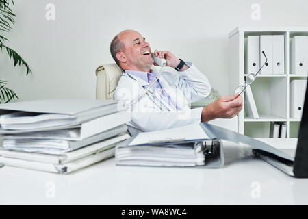 Gerne Arzt am Tisch sitzen mit Mappen und Ordner abgedeckt und Lachen bei Anrufen auf Telefon Stockfoto