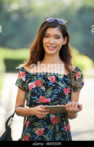 Attraktive lächelnden jungen japanischen Frau im schönen geblümten Kleid holding Tablet-PC Stockfoto