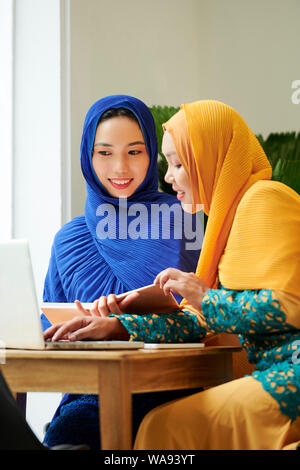 Lächelnd muslimische Frauen in traditioneller Kleidung und hijabs Buch lesen und ansehen Webinar am Laptop Stockfoto