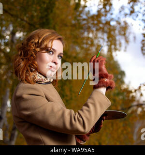 Junge schöne Frau mit Palette und Pinsel Lacke Farben Blätter an den Bäumen. Draußen im Herbst Park. Stockfoto