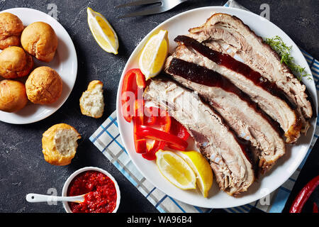 Scheiben Schweinebraten Bauch auf eine weiße Platte mit jamaikanischen gebratene Knödel und Hot Pepper Sauce in einer Schüssel auf einer konkreten Tabelle, horizontale Ansicht von Abo Stockfoto
