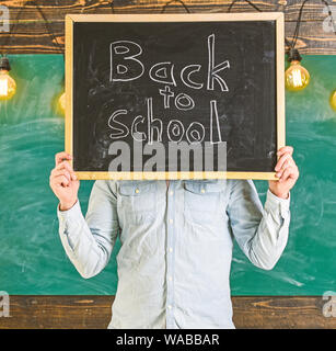 Beginn des Schuljahres Konzept. Lehrer gesichtslosen hält Tafel mit Titel zurück in die Schule. Lehrer hält Schiefertafel vor Gesicht. Mann begrüßt Studenten, schwarzes Brett für den Hintergrund. Stockfoto