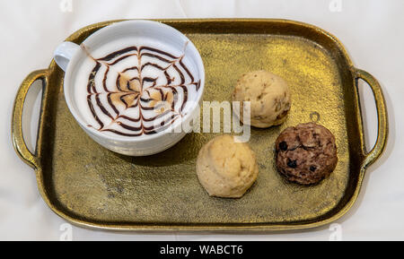 Cappuccino mit Lust auf Schokolade Design auf die Oberseite mit drei verschiedenen gebackenen Plätzchen auf Gold Tablett im Hotelzimmer auf dem Bett Stockfoto