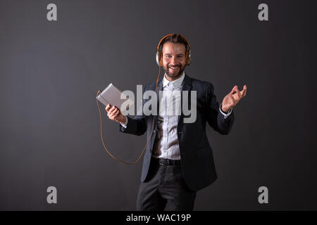 Ein lächelndes hübscher junger Geschäftsmann im Anzug mit einem Tablett und hören die Musik vor einem dunklen Hintergrund grau in einem Studio. Stockfoto