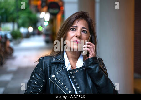 Eine ernsthafte reife Frau in schwarzem Leder Jacke zu Fuß auf der Straße und im Gespräch auf Ihrem Telefon. Stockfoto