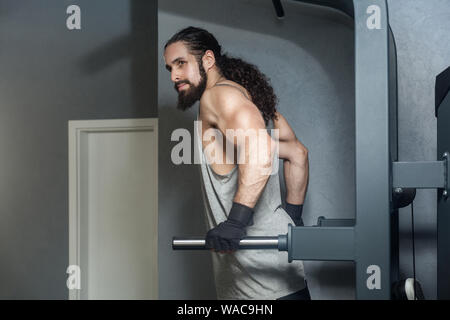 Portrait von mächtigen selbstbewussten jungen Erwachsenen starke Mann mit Langen lockigen Haar heraus in der Turnhalle arbeiten, hängen an horizontalen Balken mit Händen, Zug tut ups wi Stockfoto