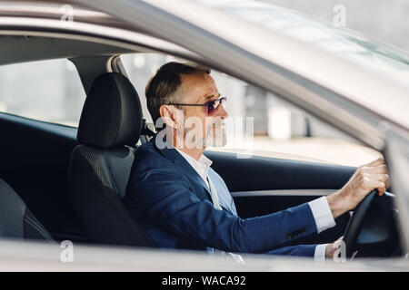 Senior Geschäftsmann, der ein Auto fährt. Seitenansicht eines Mannes, der in der formalen Verschleiß im Fahrzeug sitzen. Stockfoto