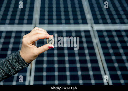 Selektiver Fokus auf Hand 1 Euro Münze und verschwommenes solar panel Zelle auf Hintergrund mit kopieren. Geld Geschäft im Bereich der erneuerbaren Energie. Stockfoto