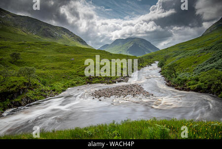Grüne Tal zwischen schönen Berghängen, Fluss mit fließendem Wasser und dramatischer Himmel in den Highlands von Schottland. Stockfoto