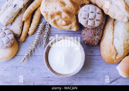 Verschiedene Arten von traditionellen Italienischen frisch gebackenes Brot und Brötchen mit Schüssel Mehl auf hölzernen Tisch Hintergrund. Rustikale Vintage Style. Ansicht von oben. Stockfoto