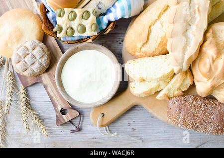 Verschiedene Arten von traditionellen Italienischen frisch gebackenes Brot und Brötchen mit Schüssel Mehl auf hölzernen Tisch Hintergrund. Rustikale Vintage Style. Ansicht von oben. Stockfoto
