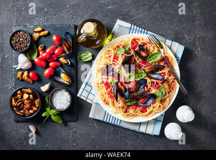 Nudeln mit Meeresfrüchten in einer würzigen Tomatensauce auf einem weißen Teller auf einen konkreten Tisch mit Zutaten auf einem schwarzen Schiefer Fach, horizontale Ansicht, close-up Stockfoto