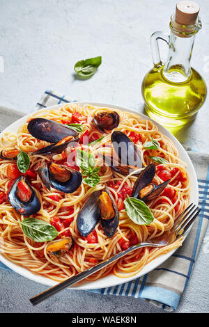 Vertikale Ansicht von Spaghetti mit Miesmuscheln in pikanter Tomatensoße und Basilikum auf einem weißen Teller auf weißem Beton Tisch mit einer Flasche Olivenöl Stockfoto