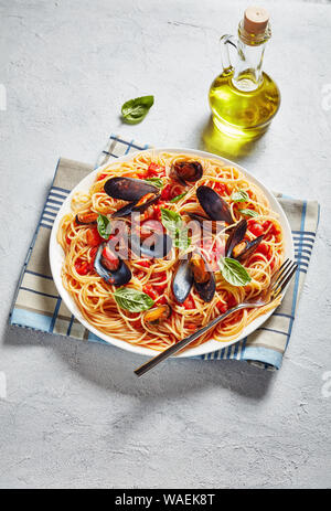 Vertikale Ansicht von Spaghetti mit Miesmuscheln in pikanter Tomatensoße und Basilikum auf einem weißen Teller auf weißem Beton Tisch mit einer Flasche Olivenöl Stockfoto