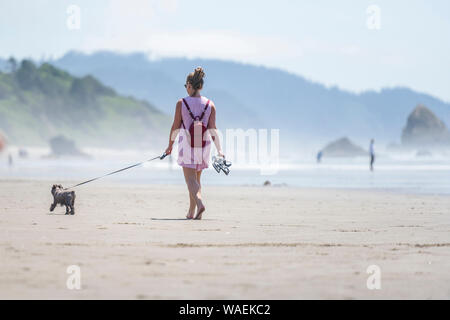 Elegante Frau in Sonnenbrille mit einem kleinen Rucksack Barfuß-Wanderungen mit einer Miniatur terrier an der Leine entlang des Pazifischen Ozeans im Nordwesten, dabei auf Stockfoto