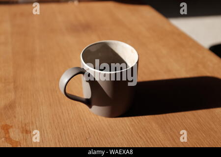 Eine halb fertige Kaffee in einem grauen Becher, warf einen langen Schatten auf einem Tisch an einem Sommermorgen. Stockfoto