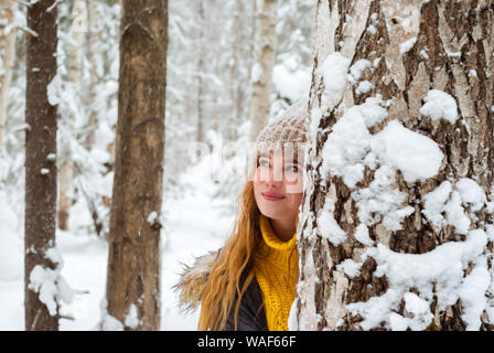 Fröhliches Mädchen in einen warmen Pullover und Strickmütze in einem Winter Park spielen verstecken sich hinter einem Baumstamm Stockfoto