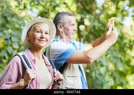 Portrait von reifer Tourist in Hut und mit Rucksack an Kamera suchen, während der Wald mit dem Mann unter Bild im Hintergrund Stockfoto