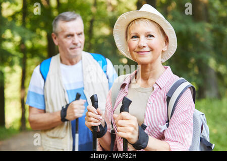 Portrait von reife Frau in Hut und mit Rucksack an Kamera suchen, während Sie Nordic Walking zusammen mit ihrem Mann in der Natur Stockfoto