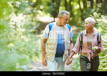 Schönes älteres Ehepaar mit Rucksäcken halten sich an den Händen und reden miteinander während ihrer Wanderung im Wald im sonnigen Sommertag Stockfoto
