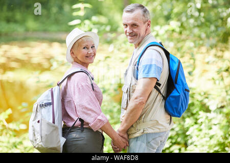 Portrait von Senior paar Wanderer mit Rucksack auf dem Rücken stehend, Hände halten und Lächeln auf die Kamera in der Natur im Wald Stockfoto