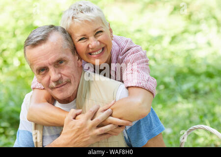 Portrait von Happy reife Frau ihrem Mann umarmt, während hinter seinem Rücken stehend und sie lächelnd in die Kamera im Freien im Sommer Tag Stockfoto