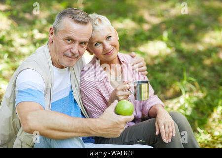 Portrait von Reifes Paar sitzt auf dem Gras und umarmen einander beim Tee trinken und Obst essen im Freien Stockfoto