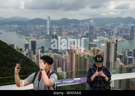 Hongkong, China. 20 Aug, 2019. Zwei Touristen stehen auf dem Victoria Peak vor die Skyline von Hong Kong und auf ihren Smartphones. Anders als in den Wochen vor, die Proteste am Wochenende nicht zu erheblichen Unruhen führen. Die Demonstranten nicht Barrikaden zu errichten. Die Polizei verzichtete mit Gas, das als ein Zeichen der Entspannung angesehen wurde. Credit: Gregor Fischer/dpa/Alamy leben Nachrichten Stockfoto