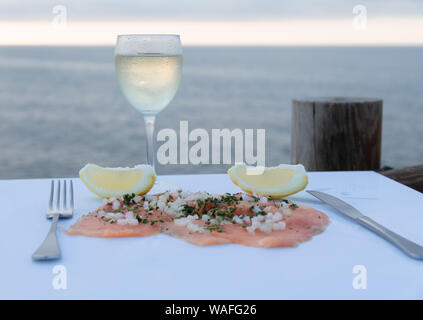 Luxuriöse geräucherter Lachs filet mit Sellerie, Zwiebel, Scheiben von Reife Zitrone und einem Glas Wein auf einem weißen Tisch - Das Meer im Hintergrund Stockfoto