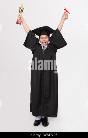 Junge im Abschlusskleid zeigt einen Abschluss mit Trophäe Stockfoto