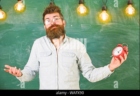 Lehrer in Brillen hält Wecker. Zeitplan und Regime Konzept. Mann mit Bart und Schnurrbart auf verwirrten Gesichtsausdruck im Klassenzimmer. Bärtige hipster hält, schwarzes Brett für den Hintergrund. Stockfoto
