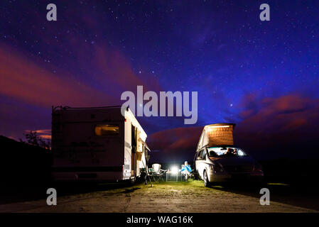 Wohnmobile sind auf einem Strand bei Nacht unter den Sternen geparkt. Ein Mann mit zwei Camping Camping Vans genießen die Entspannung am Abend unter thew Starry Night Stockfoto