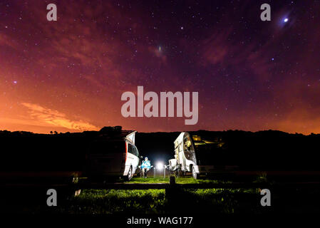 Wohnmobile sind auf einem Strand bei Nacht unter den Sternen geparkt. Ein Mann mit zwei Camping Camping Vans genießen die Entspannung am Abend unter thew Starry Night Stockfoto