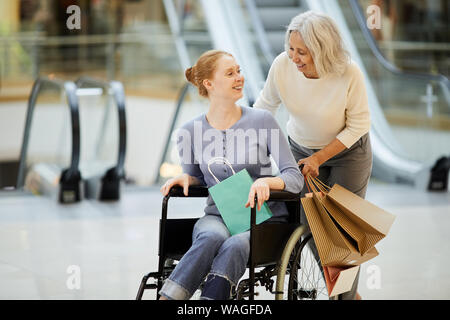 Ältere Mutter mit Tüten lachen und sprechen mit ihrer Tochter während ihrer Durchführung im Rollstuhl im Einkaufszentrum Stockfoto