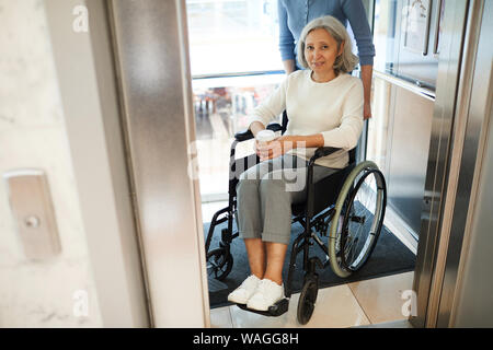 Portrait von älteren weiblichen Patienten Sitzen im Rollstuhl mit Tasse Kaffee und die Kamera während der Krankenschwester helfen, aus dem Aufzug zu erhalten Stockfoto