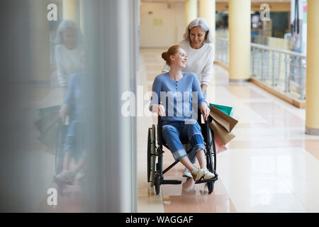 Gerne ältere Mutter mit Tüten ihre behinderte Tochter, die im Rollstuhl Sie reden und lächeln in der Mall Stockfoto