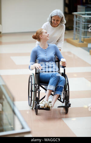 Behinderte junge Frau im Rollstuhl lächelnd zu ihrem älteren Mutter, während Sie zu Fuß entlang der Shopping Mall Stockfoto