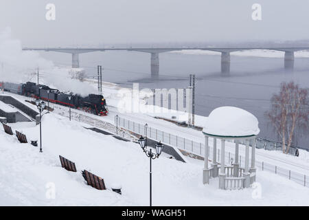 Winter verschneite Landschaft mit Dampflokomotive in Bewegung entlang der Ufer mit einem Vintage Rotunde und Automobil Bridge im Hintergrund Stockfoto