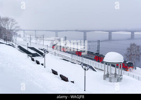 Winter verschneite Landschaft mit Elektrischer Triebzug Zug in Bewegung entlang der Ufer mit einem Vintage Rotunde und Automobil Bridge im Hintergrund Stockfoto