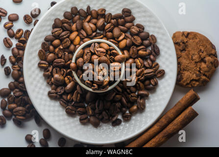 Weiß Kaffeetasse mit gerösteten Kaffeebohnen, Zimt und Keksen auf isolierten weißen Hintergrund von oben. Kaffeebohnen auf Platte und in der Tasse. Stockfoto