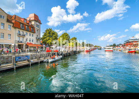 Touristen genießen Geschäfte, Boote und Cafes an der Promenade am Alten Strom Kanal in der Stadt Warnemünde, Rostock an der nördlichen Küste von Deutschland Stockfoto