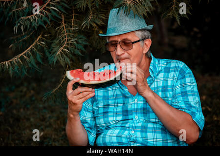 Gerne älterer Mann mit Hut Essen saftige Wassermelone im Park Stockfoto