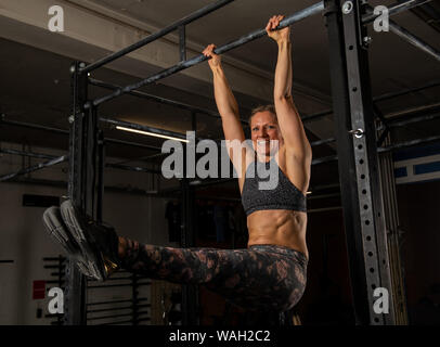 Eine schöne blonde Frau mittleren Alters mit Sixpack ist die Übung l sitzen auf der horizontalen Leiste. Funktionelle Fitness Training in einem Fitnessstudio. Stockfoto