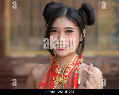 Oung Thai chinesische Schönheit mit Chun Li doppel Haar Brötchen trägt einen eleganten rot-goldenen Chinesische Kleid (CHEONGSAM) und posiert für die Kamera (finger Herz). Stockfoto