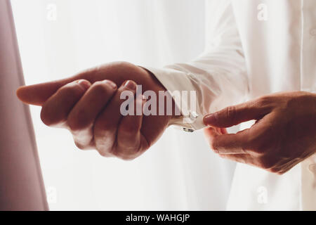 Der Mann befestigt ist, und die Manschetten des Hemdes. Am Morgen des Bräutigams. Hände von Hochzeit Bräutigam Knopfverschluß, sein weißes Hemd. Der männliche Hände auf dem Hintergrund einer Stockfoto