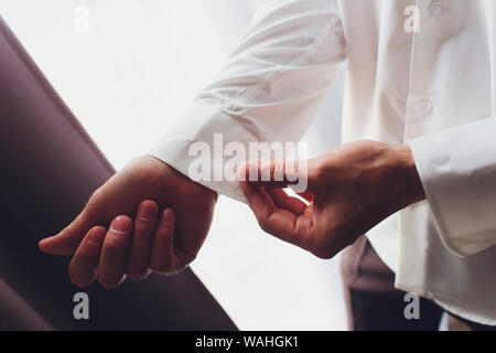 Der Mann befestigt ist, und die Manschetten des Hemdes. Am Morgen des Bräutigams. Hände von Hochzeit Bräutigam Knopfverschluß, sein weißes Hemd. Der männliche Hände auf dem Hintergrund einer Stockfoto