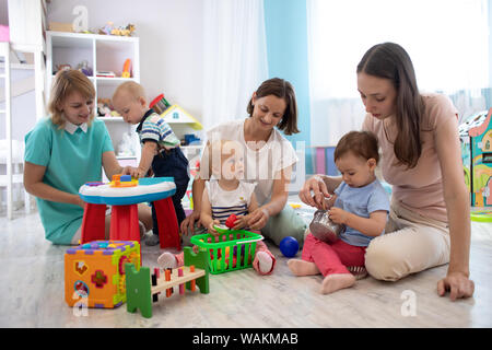 Adorable Kleinkinder spielen mit farbenfrohen Spielzeuge und Mütter im Kinderzimmer. Baumschule Babys spielen mit Erwachsenen in der Kindertagesstätte Stockfoto