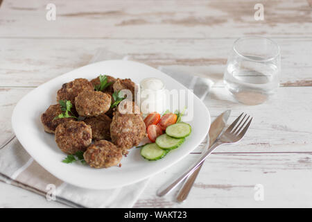 Saftige köstliche Fleisch Schnitzel in eine weiße Platte auf hellem Holztisch. Metballs gebraten serviert mit Tomaten und Gurken und Sauerrahm. Stockfoto