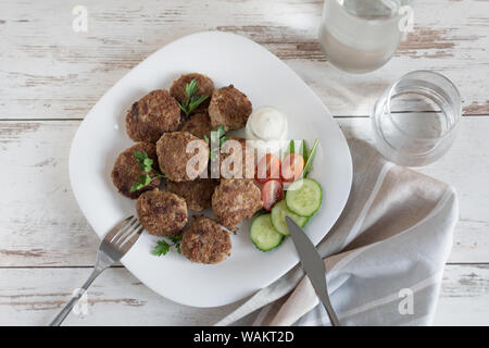 Saftige köstliche Fleisch Schnitzel in eine weiße Platte auf hellem Holztisch. Metballs gebraten serviert mit Tomaten und Gurken und Sauerrahm. Stockfoto